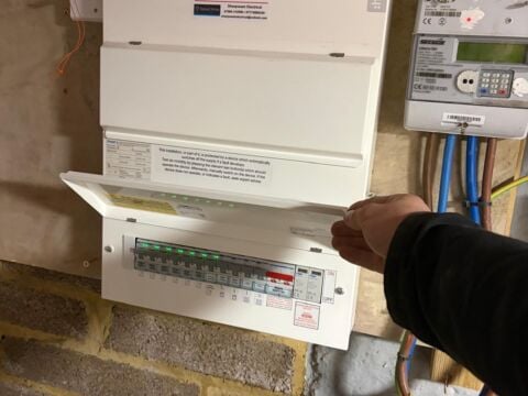 An electrician inspects an open consumer unit and fuse box installation mounted on an interior wall, with visible circuit breakers, electrical wiring