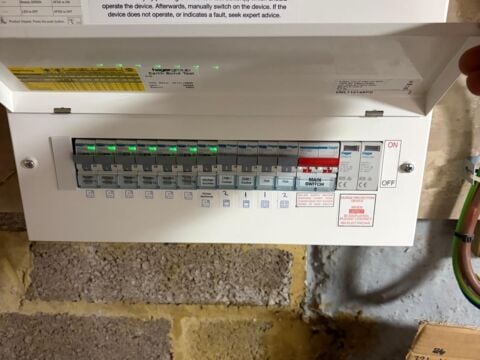 White consumer unit installed on a painted brick wall, showing multiple circuit breakers with green indicators, a main switch, and RCD protection