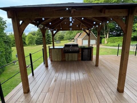 Outdoor timber pergola with pitched roof covering a wooden deck area, featuring an integrated outdoor kitchen unit with barbecue and storage