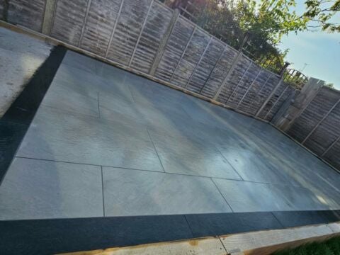 Large format grey porcelain patio slabs laid in a residential garden with timber edging frame and horizontal wooden fence panels along the boundary