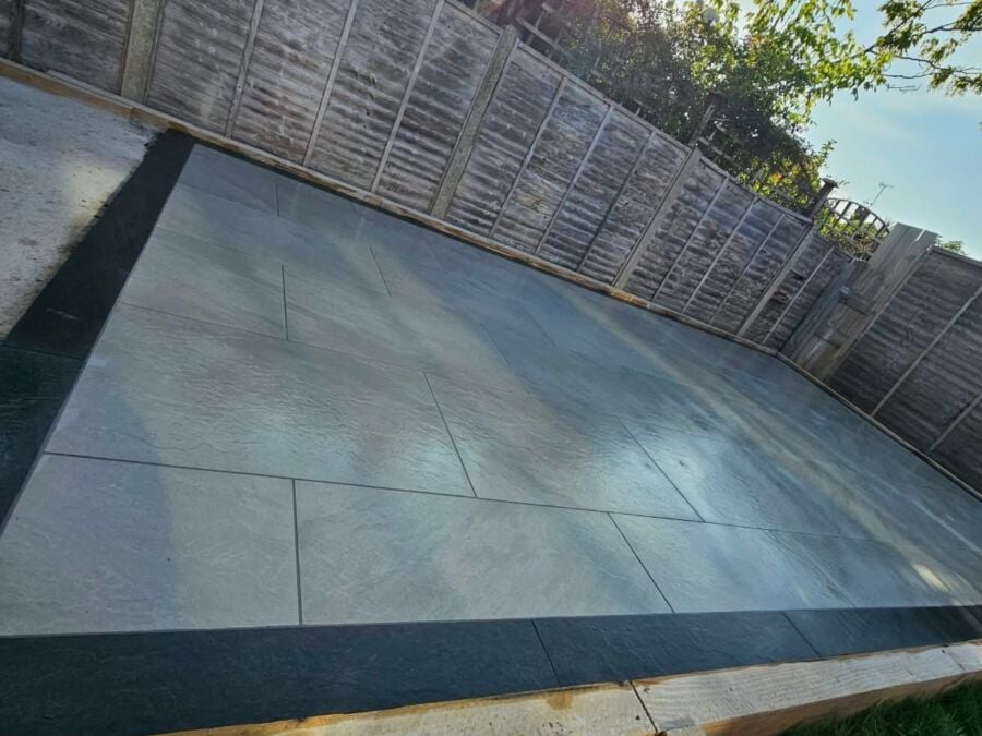 Large format grey porcelain patio slabs laid in a residential garden with timber edging frame and horizontal wooden fence panels along the boundary