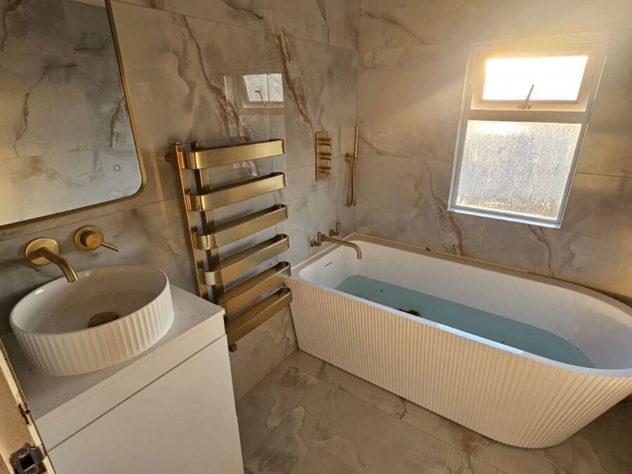 Contemporary bathroom installation featuring marble effect wall and floor tiles, freestanding ribbed bath with brushed brass fittings