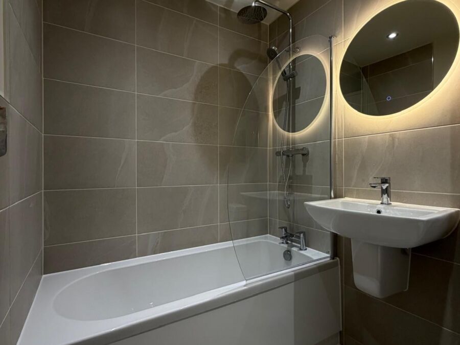 Bathroom with bath, shower screen, grey tiles and round backlit mirror