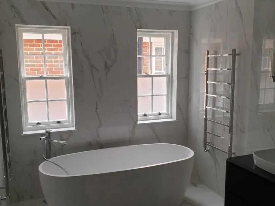 Modern bathroom featuring a freestanding white oval bathtub positioned beneath two sash windows, with large format marble effect wall tiles